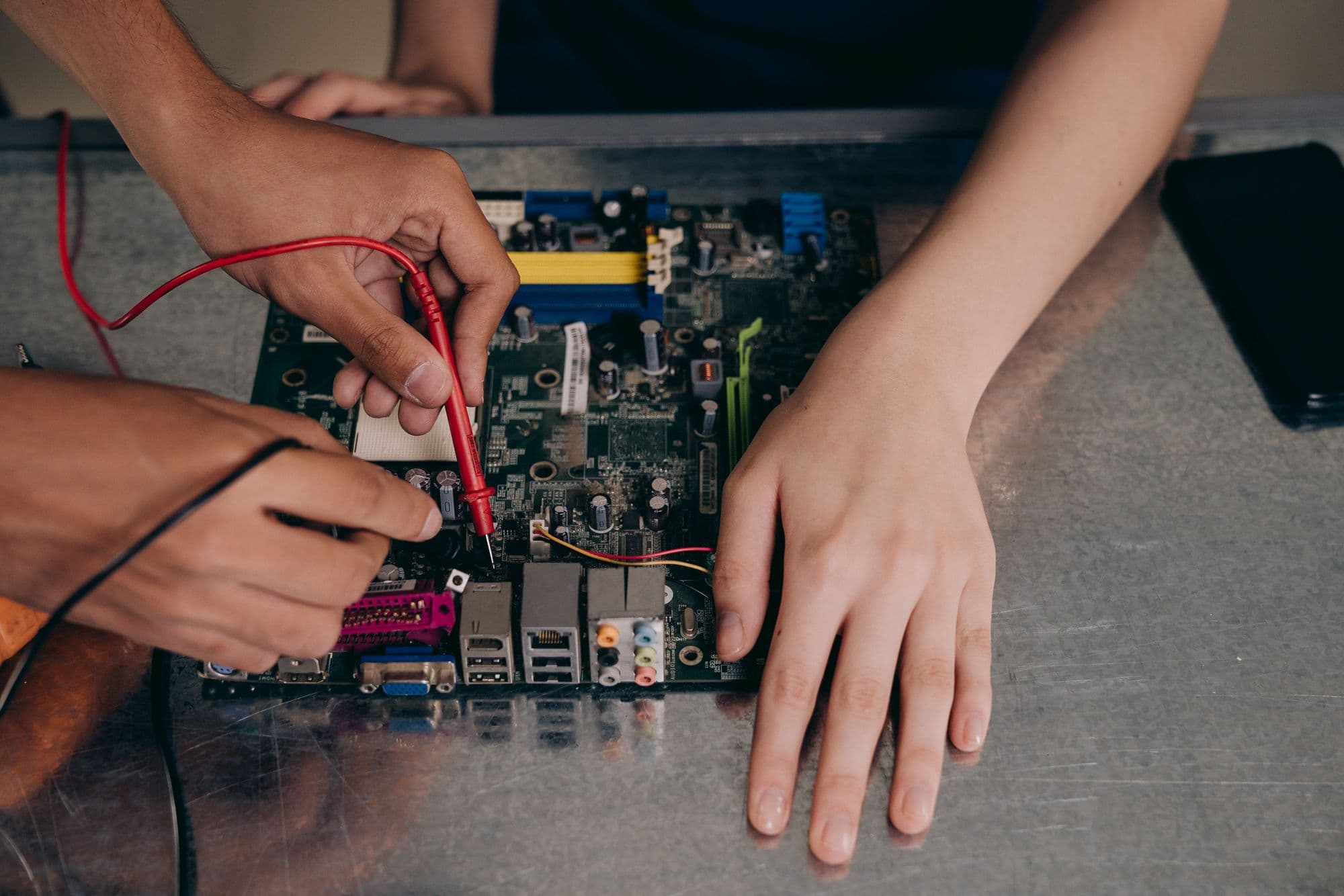 Hands testing a desktop motherboard with multimeter probes on a steel workbench