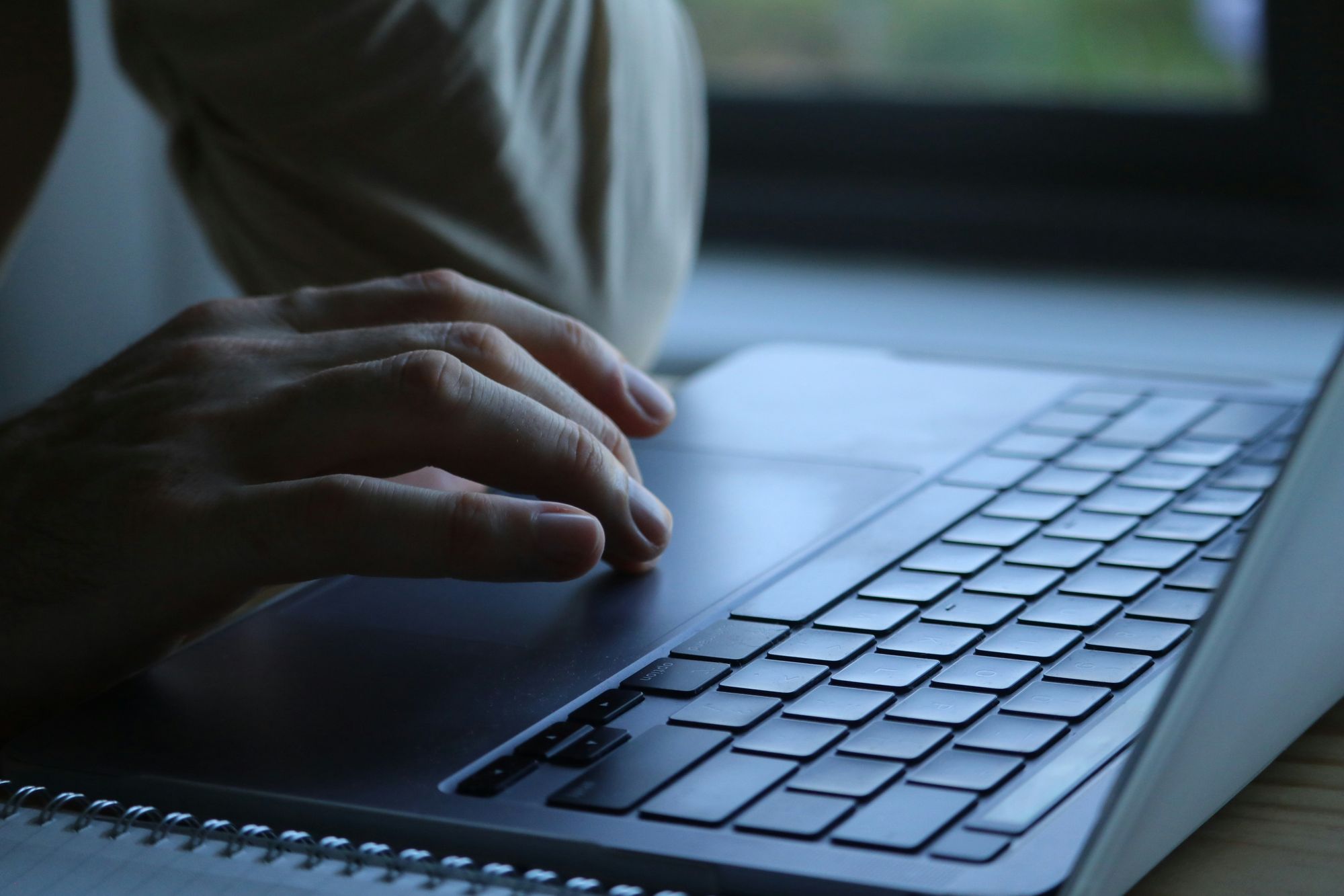 A hand on the trackpad of a dark grey laptop beside a window